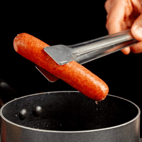 A caucasian woman lifting up a single hot dog out of boiling water using a metal tong.