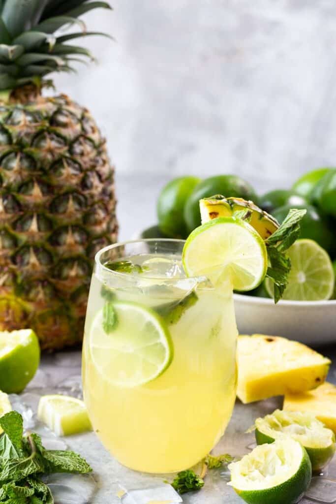 glass of pineapple mojito with pineapple and limes in background