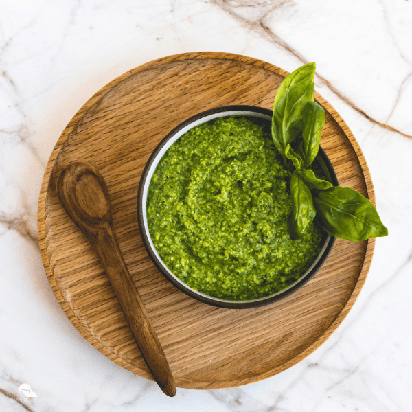 A top shot of pesto in a bowl, on top of a circular wooden tray. A wooden spoon is also included in the image.