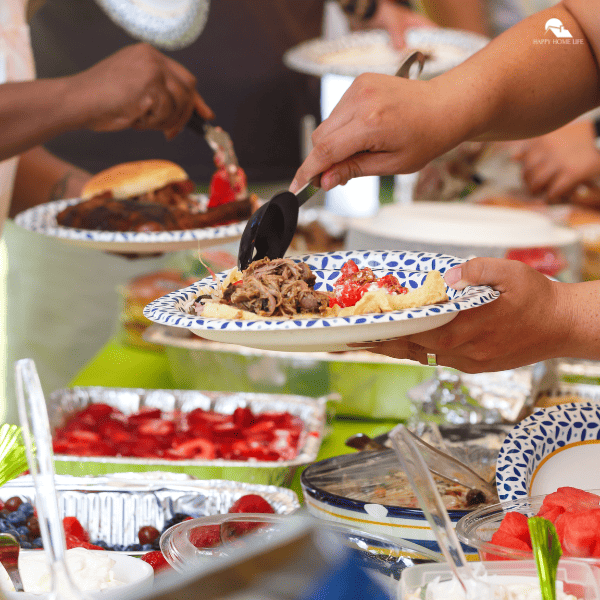 An image of a potluck, with hands putting food in their paper plates. Different foods are also seen in the background.