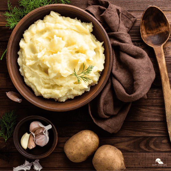 An image of mashed potatoes in a wooden bowl, with garlic cloves, potatoes, and a wooden spoon surrounding it.