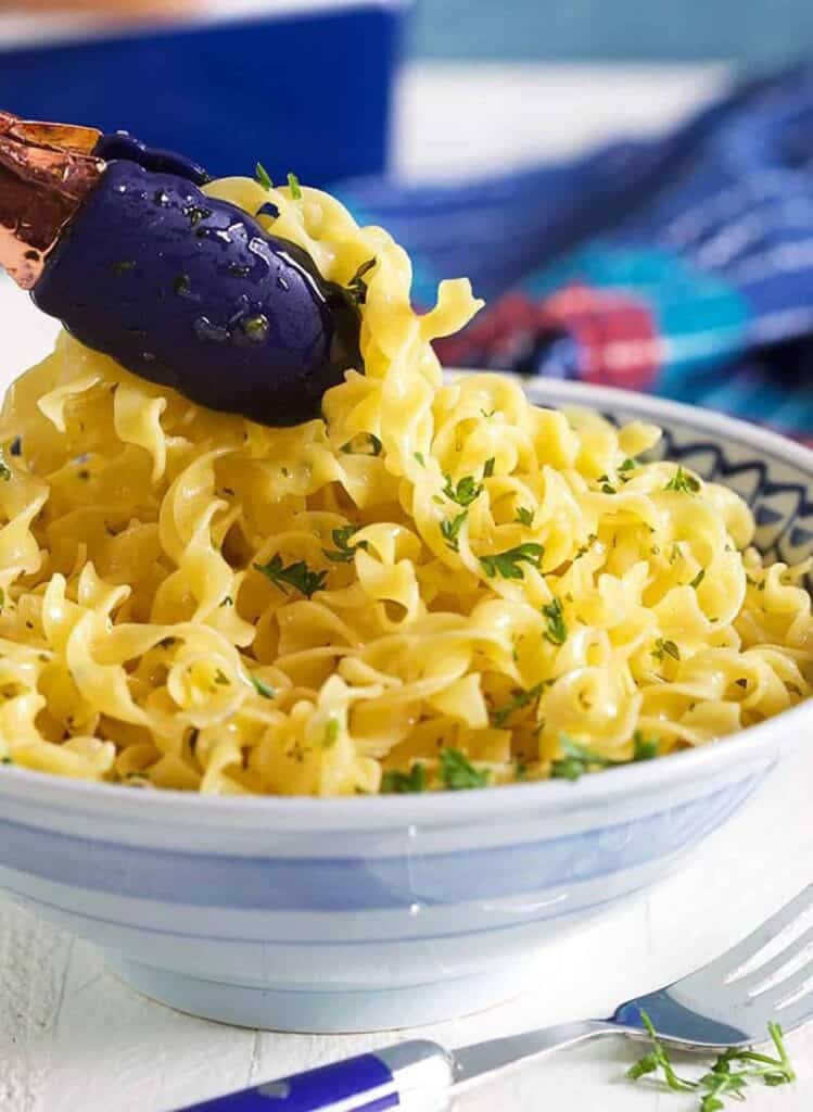 tongs holding buttery egg noodles over blue and white bowl
