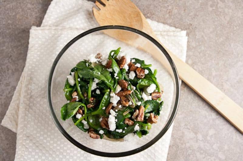 wilted spinach salad in clear bowl on top of white cloth, wooden spoon off to side