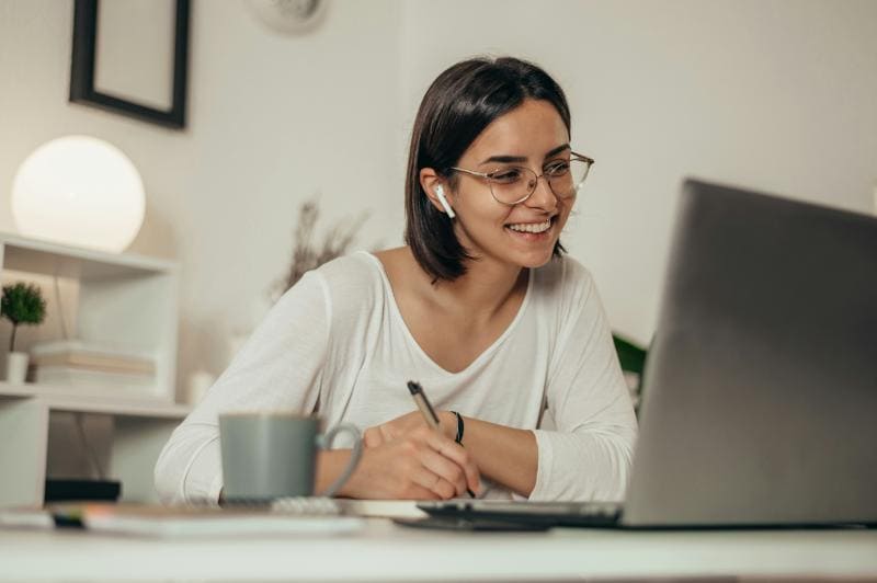 woman with glasses and earbuds in using laptop and taking notes in home office