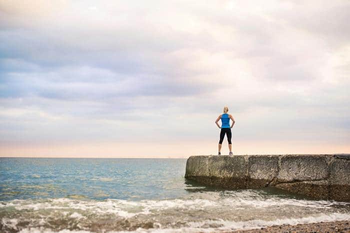 woman in exercise clothes standing triumphantly on a rock near the ocean