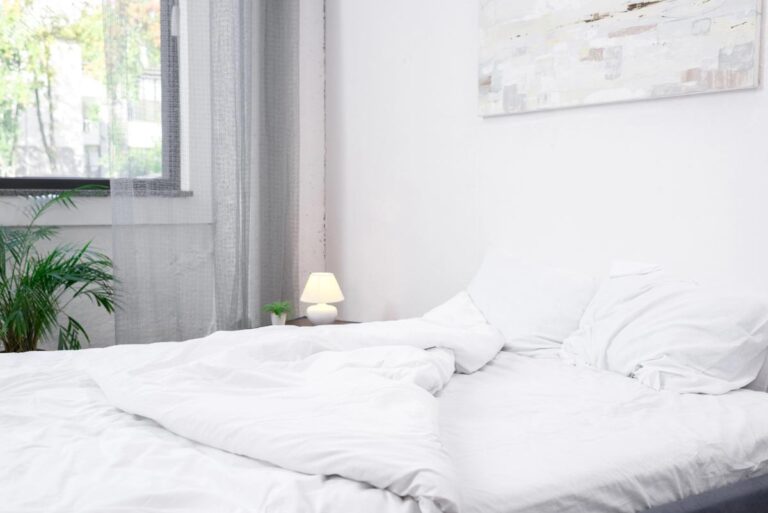 an empty bed with white bedding near a window