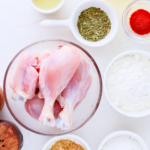 Raw ingredients for crispy fried chicken including chicken drumsticks, flour, cornstarch, paprika, mixed herbs, garlic powder, and oil arranged on a white surface.