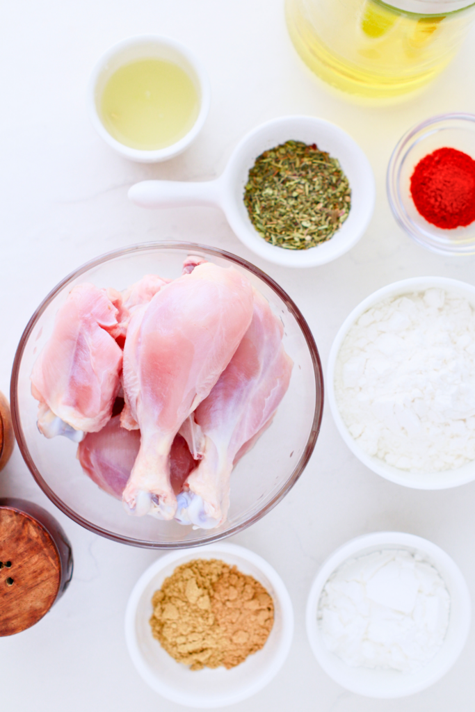 Raw ingredients for crispy fried chicken including chicken drumsticks, flour, cornstarch, paprika, mixed herbs, garlic powder, and oil arranged on a white surface.