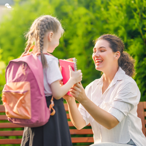 A smiling mother kneels in front of her young daughter wearing a backpack, handing her a notebook before school.