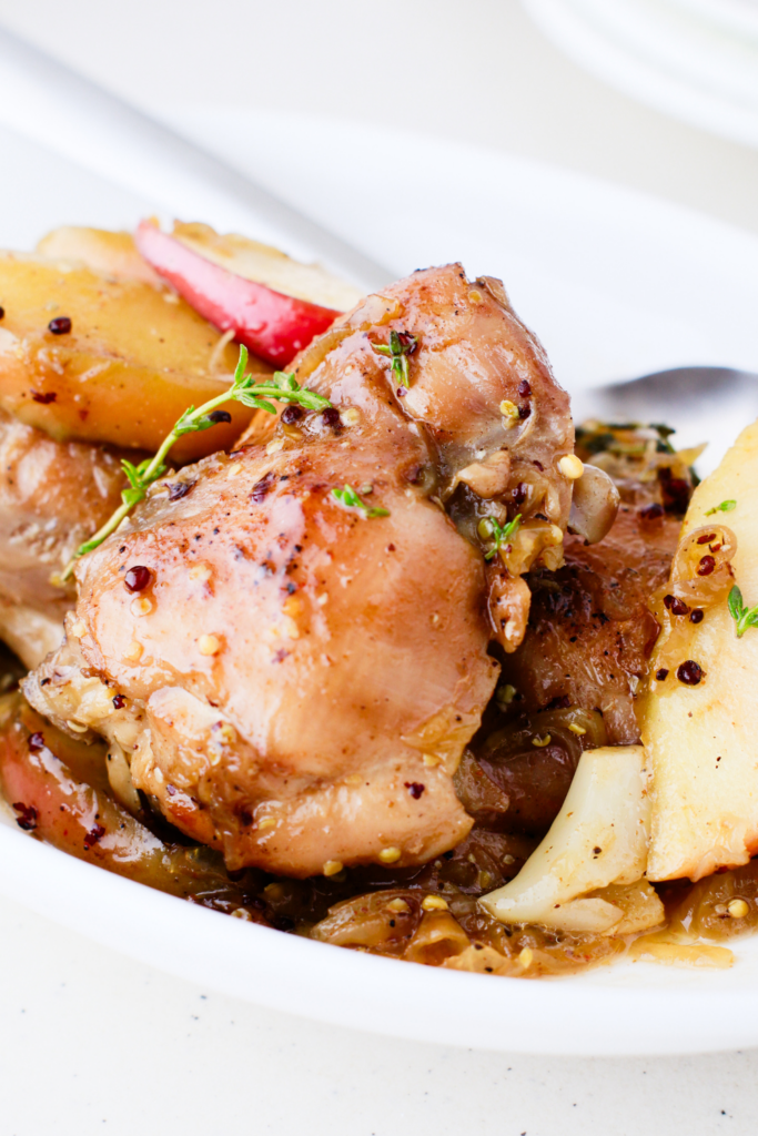 Apple cider chicken thighs served in a white dish, surrounded by caramelized apples, onions, and fresh thyme.